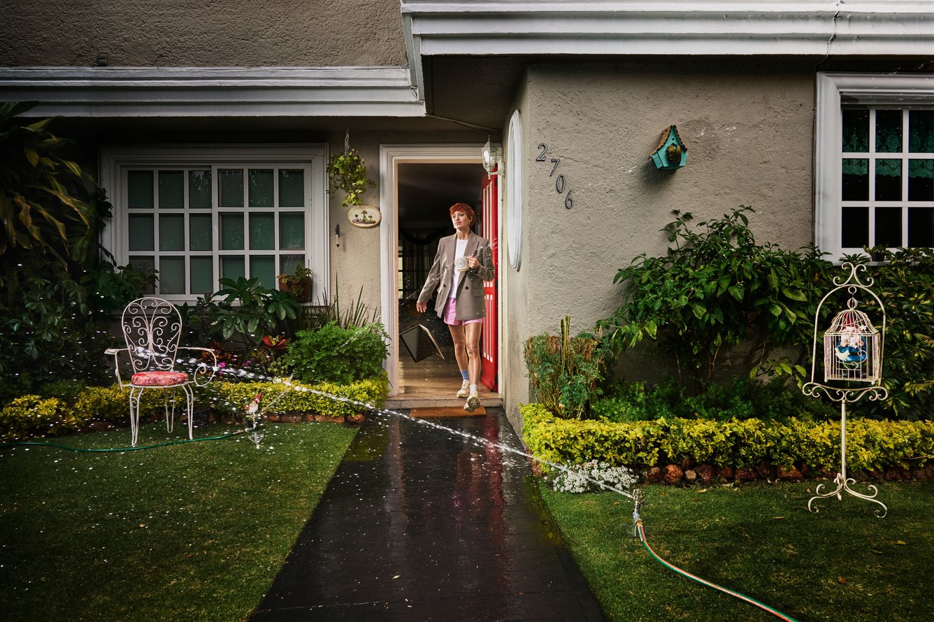 A woman wearing a blazer, shorts, and athletic shoes steps out her house's front door into a yard with a sprinkler spraying water and green grass.